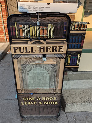 An old newspaper stand with books and library painted on it. Text on the stand reads, "Take a book, leave a book."