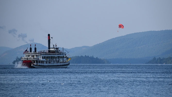 A steamship with a paddlewheel crossing a blue lake with blue mountains surrounding it