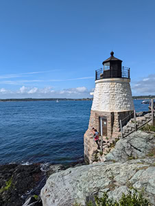 A small white lighthouse with a black top is nestled between low cliffs and the blue bay