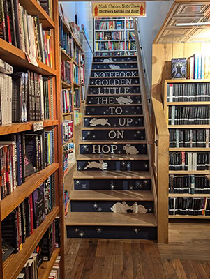 A stairwell surrounded by books. The stair risers are painted with white bunnes and the words "Hop on up to the Little Golden Notebook."