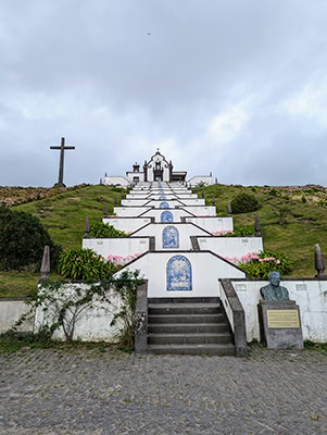 Stairs with pink flower and white landings and tiles depicting the life of Jesus climb a hillside to a white chapel and a cross