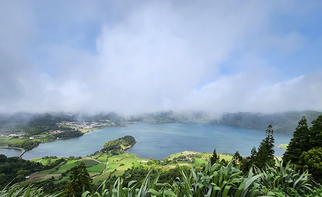 Looking over lush green vegetation, there's a blue lake below in a volcanic cauldron with wisps of white fog passing through