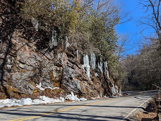 Icicles hang from a rocky cliff beside a curving road on a blue-sky day