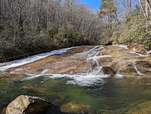 A "natural water slide" cascades down a gently sloping rock face with ice and snow along the edges and clear green water in the plunge pool