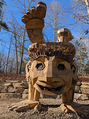 A larger-than-life troll made of wood smiles while doing a handstand in the winter woods