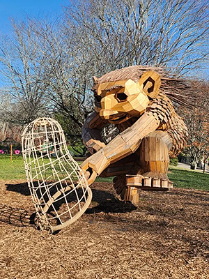 A larger-than-life troll made of wood brings a wooden net to the ground as if capturing a scientific specimen